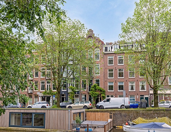 Appartementengebouwen aan een Amsterdamse kade met bomen, geparkeerde auto’s en uitzicht over het water